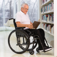 A senior man sits smiling in his Move wheelchair. He's reading a book in a brightly lit space in front of a shelf of books. There are windows behind him with a view to leafless trees. The space is painted white with pillars visible in the distance. thumbnail